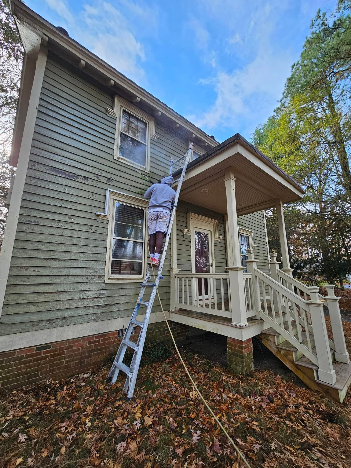 Crew member on a ladder washing the failing painted siding of a historic farmhouse before repainting