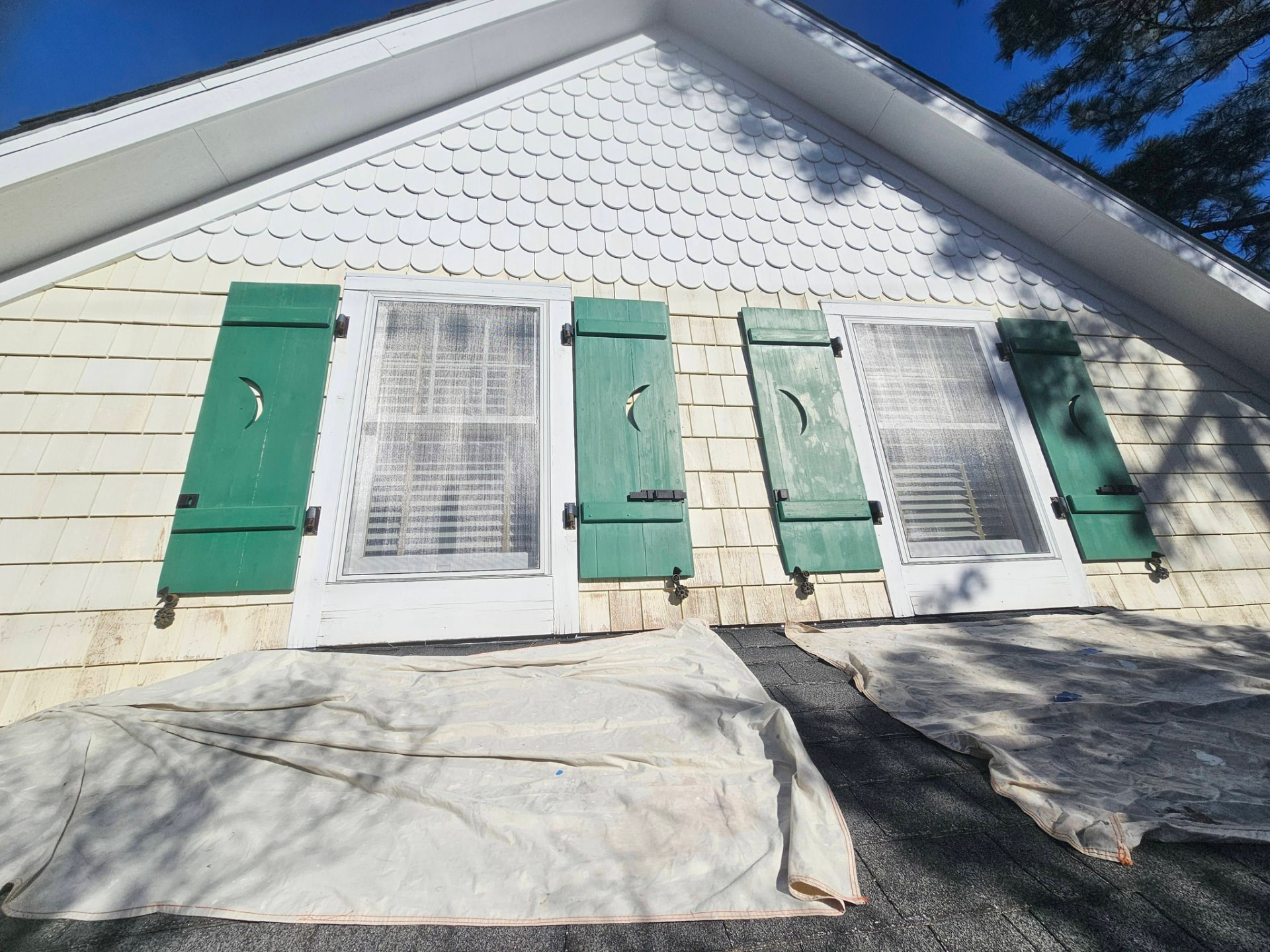 Upper gable with weathered, chalky green moon-cut shutters and dingy shingles, drop cloths on the roof